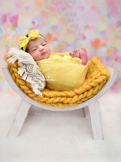 This baby girl is resting on a curved wooden bench, wrapped in a sunny yellow swaddle that contrasts beautifully with the pastel background.