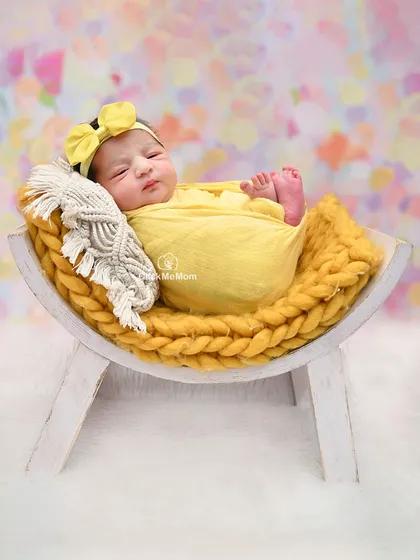 This baby girl is resting on a curved wooden bench, wrapped in a sunny yellow swaddle that contrasts beautifully with the pastel background.