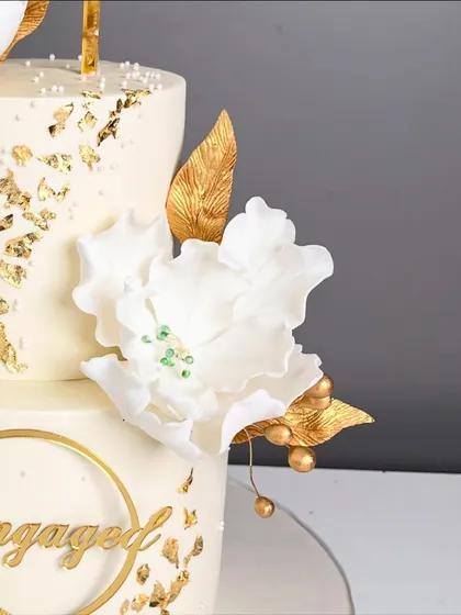 A close-up of a dreamy engagement cake, highlighting a delicate, handcrafted white sugar flower with gold accents and a sprinkle of gold leaf.