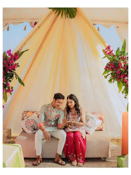 The couple sits together under a canopy, admiring the bride's freshly applied Mehendi.