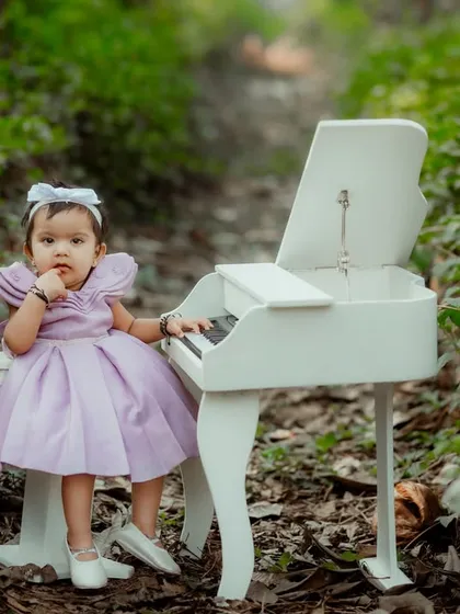 A whimsical portrait of a baby girl in a lovely lavender dress, sitting at a miniature piano in an enchanted forest-like setting.