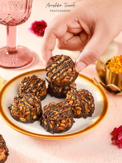 A hand reaching for a chocolate-drizzled nut cluster. The festive colours in the background and the action in the shot make it feel like a moment from a real Diwali party.