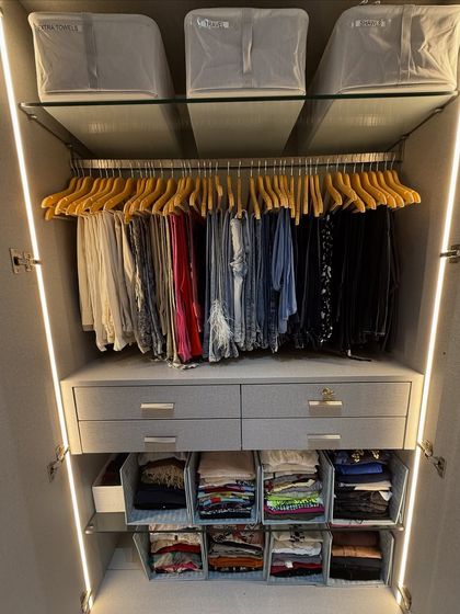 This closet shows a system where trousers are hung on uniform hangers and folded items are categorized in clear bins below. The top shelf is used for bulky items stored in matching fabric boxes.