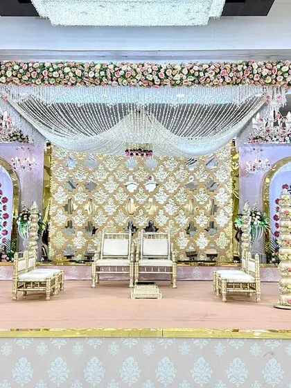 This wedding mandap combines intricate mirror work with a soft pastel theme. The backdrop features a central gold mirror panel flanked by arches with delicate floral paintings. The entire structure is crowned with a thick border of pastel roses and hanging crystal strings.