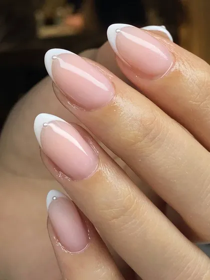 A modern twist on a classic. This elegant French manicure on almond-shaped nails is elevated with a tiny, delicate pearl accent at the base of each tip, adding a touch of sophisticated detail.