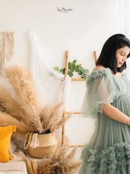 The soft green gown blends beautifully with the natural light and bohemian decor of the studio, creating a peaceful and dreamy portrait by the window.