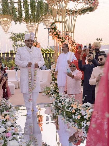 The groom awaits his bride on the mirrored aisle, with the grand floral mandap in the background, creating a moment full of anticipation and beauty.