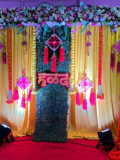 A classic Haldi ceremony backdrop with yellow curtains and a central green panel. The stage is adorned with marigold garlands and traditional decorative pieces.