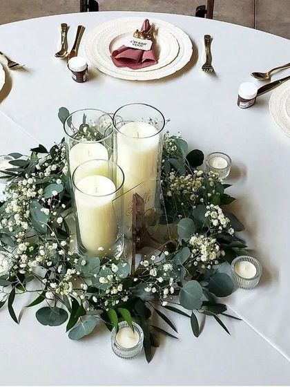 A simple yet elegant centerpiece design. A wreath of baby's breath and greenery surrounds glass hurricane lamps with pillar candles, perfect for a minimalist and romantic table setting.