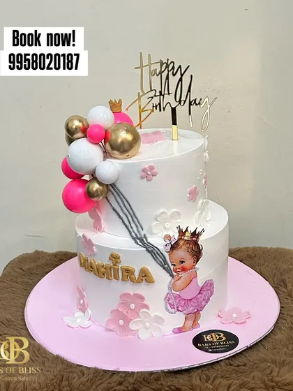 Celebrate a first birthday with this adorable two-tiered princess-themed cake. It features a cute baby princess graphic, balloon decorations, and personalized name, all in a lovely pink and white color scheme.