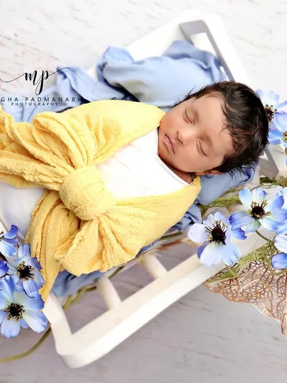 A baby wrapped in a yellow bow, sleeping on a white diwan surrounded by blue flowers.