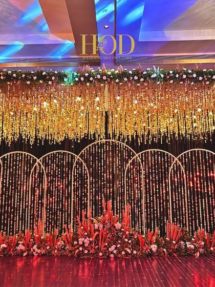 A glamorous sangeet or reception stage with a cascading gold ceiling, arched backdrops with string lights, and a base of vibrant red flowers.