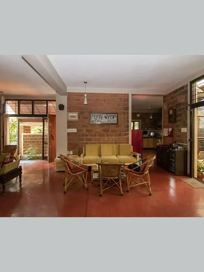 The main living area, designed as a modern 'mandapa', with cane furniture, red oxide flooring, and sliding doors opening to a garden.