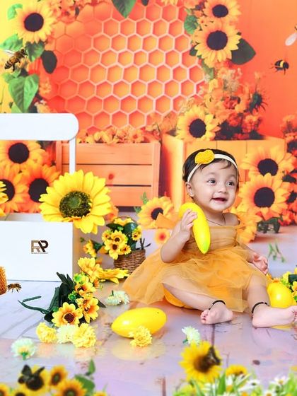 A baby girl in a yellow dress sits in a vibrant sunflower and honeybee-themed setup. The bright colors and cheerful props create a sunny and joyful atmosphere.
