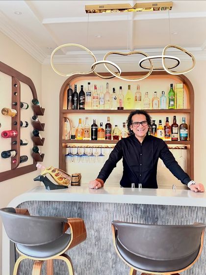 A photo of me at the finished home bar, showcasing the elegant design with its curved wooden shelving and modern pendant light.