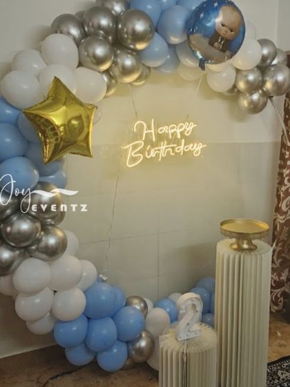 This is another take on the balloon ring decoration, perfect for a home setting. The arch of blue, white, and silver balloons is accented with a star foil balloon and a 'Happy Birthday' neon sign, with a small Boss Baby balloon detail.