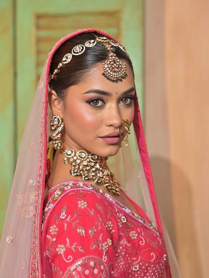A stunning close-up of a model, her face partially veiled by a pink dupatta, showcasing a heavy Kundan choker, maang tikka, and nath.