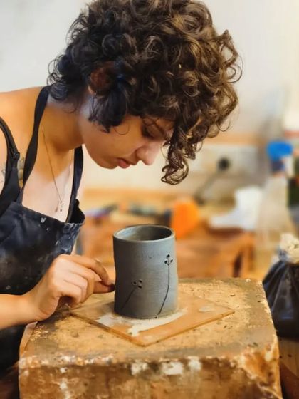 A student carefully carves delicate dandelion designs onto her hand-built pot. This technique, known as sgraffito, involves scratching through a layer of color to reveal the clay body underneath.