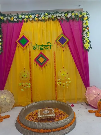 A beautiful Mehandi setup for a home ceremony. The backdrop of yellow and pink drapes is decorated with a floral border and traditional hangings, with a large urli for the rituals.