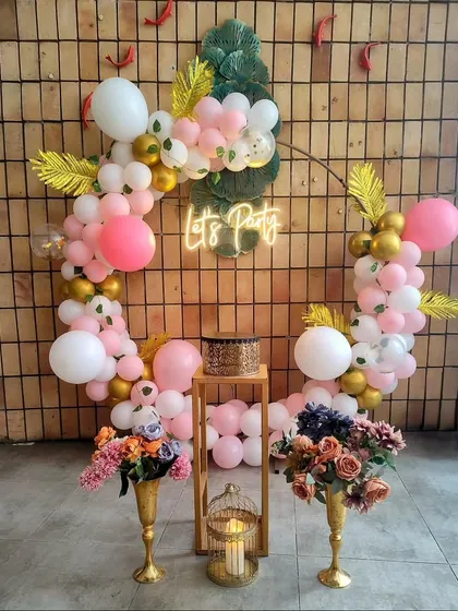 A fun and tropical "Let's Party" backdrop. The circular frame is decorated with pink, white, and gold balloons and accented with gold-painted palm leaves and a neon sign.