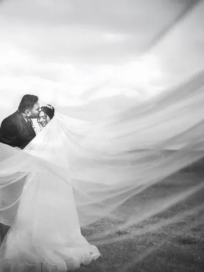 A classic black and white portrait using the bride's long veil to create a sense of movement and intimacy. The sweeping fabric against the open landscape adds a layer of drama and romance to this tender moment.