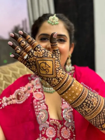 A close-up of the back-hand bridal mehendi, showcasing the personalized initial 'S' and the detailed work on the arm.