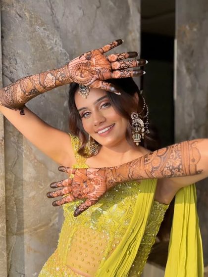 A happy bride in a lime green outfit, her hands crossed to show off her beautiful full-arm mehandi.