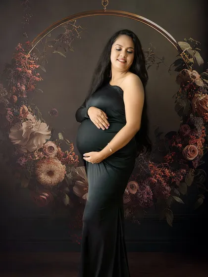 A striking solo maternity portrait featuring a mom-to-be in a sleek black gown, framed by a beautiful floral hoop. This shows a more modern, moody, and elegant studio setup.