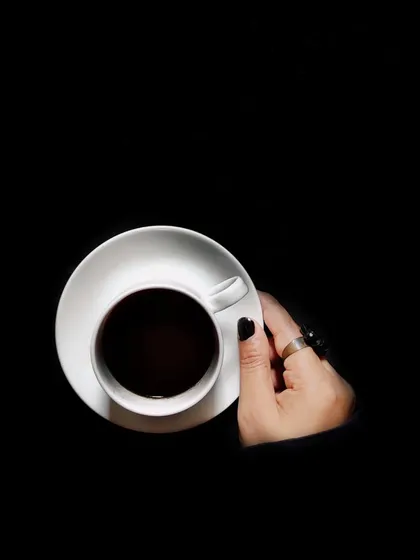 A minimalist, top-down view of a hand holding a cup of black coffee against a pure black background, creating a bold and graphic image.