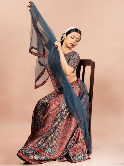 A model seated gracefully in a printed lehenga, using the sheer dupatta to create a sense of movement and elegance. This is a beautiful shot for an ethnic wear catalogue.