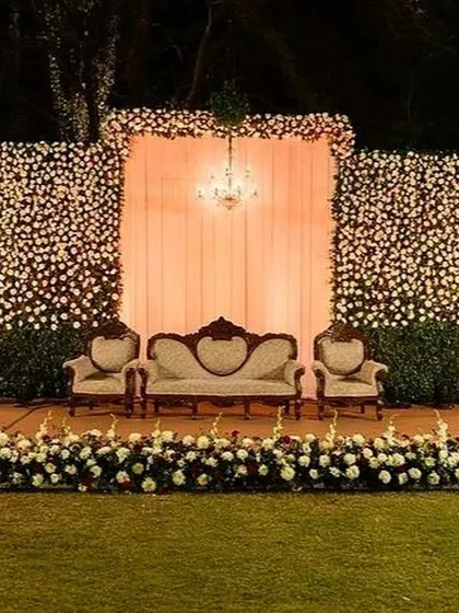 A beautiful Sangeet stage featuring a massive wall of white and red flowers. The vintage-style seating and a central chandelier add a touch of classic elegance to the outdoor lawn setting.