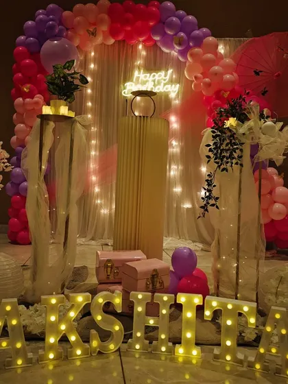 A vibrant and stylish 12th birthday celebration. The decor features a pink and purple balloon arch, a 'Happy Birthday' neon sign against a light curtain, and elegant props like cherry blossom arrangements and marquee letters spelling out the birthday girl's name.