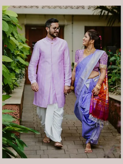 A Maharashtrian couple walks hand-in-hand through a lush green pathway, sharing a look of love and companionship in their traditional wedding attire.