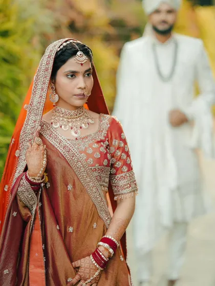 A creative shot of the bride in focus with the groom blurred in the background, building anticipation for the moment they meet at the mandap.