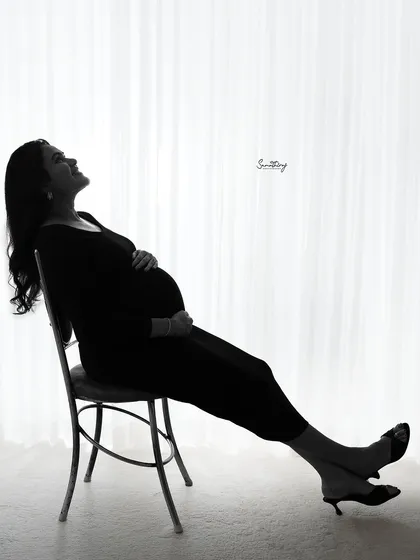 A serene and contemplative silhouette of a mother-to-be seated by a window. The bright backlighting creates a peaceful and artistic portrait that emphasizes her beautiful form.