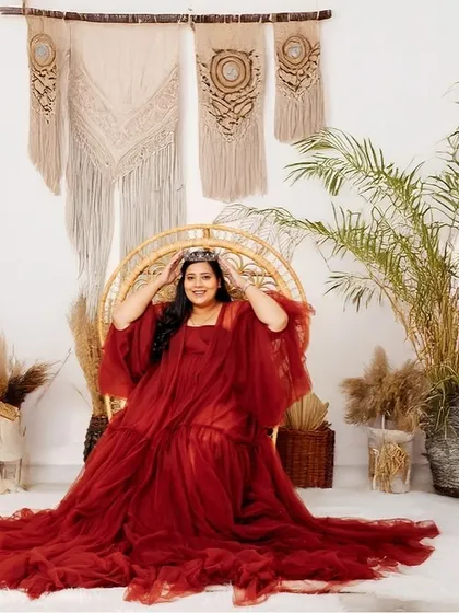A regal portrait of a mother-to-be in a stunning red gown, seated on a wicker peacock chair. The bohemian-themed studio setup includes macrame wall hangings and pampas grass, creating a warm, stylish, and earthy vibe.