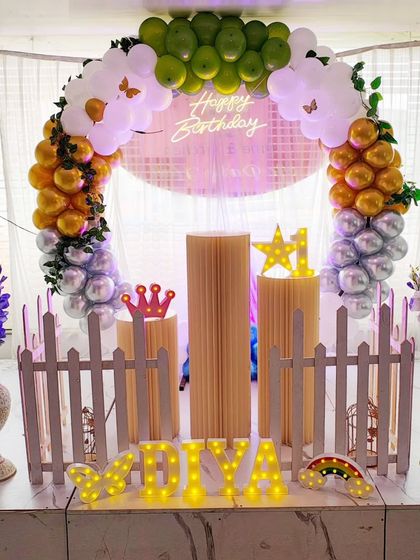 A first birthday decoration with a pastel-colored balloon arch and a translucent backdrop. The setup is accented with a neon sign, light-up marquee letters, and flower vases.