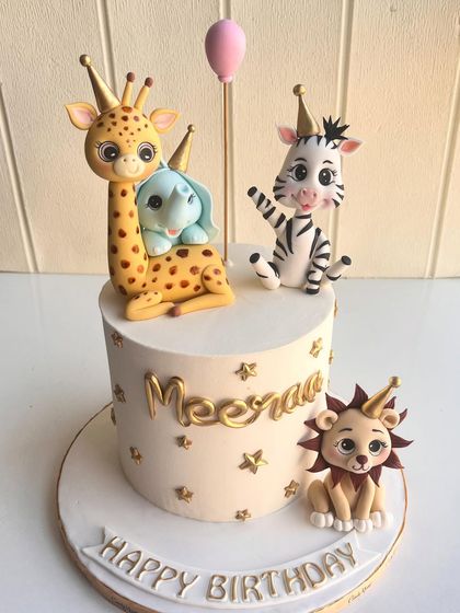 A wild jungle party on a cake. This design features adorable, hand-sculpted safari animals like a giraffe, zebra, and lion, all wearing party hats and surrounded by golden stars for a festive birthday celebration.