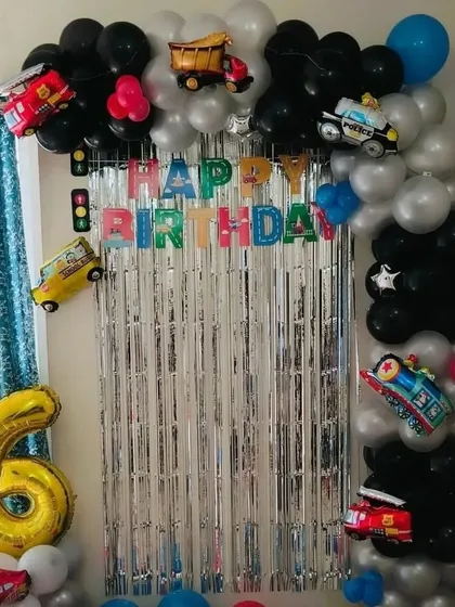 A vehicle-themed birthday decoration for a 6th birthday. The setup includes a balloon arch decorated with foil balloons of a police car, fire truck, and ambulance.