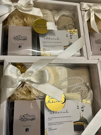 A batch of Umrah return gifts, elegantly packed in white boxes with satin ribbons and a gold "Umrah Mubarak" tag.
