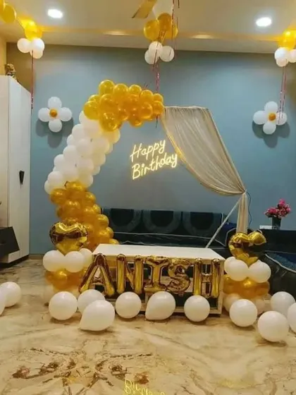 A cozy and elegant birthday setup in a living room. A white and gold balloon arch frames a small tent, with the name 'ANISH' spelled out in foil balloons.