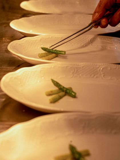 A close-up of a chef's hand using tweezers to delicately place asparagus on a plate.