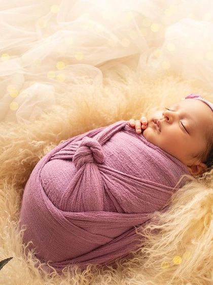 A newborn baby sleeps on a bed of soft fur, wrapped in lavender with dreamy lights creating a magical bokeh effect.