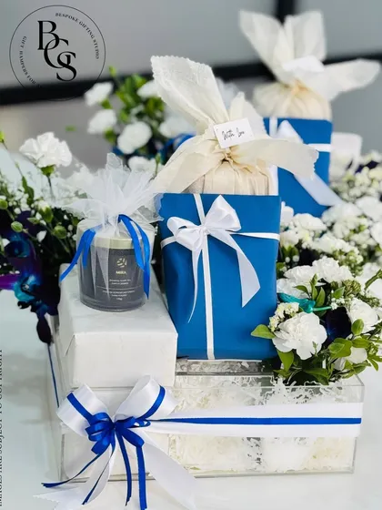 A stunning multi-layered gift arrangement in a blue and white color scheme. It features gifts wrapped in different papers and fabrics, all tied together with matching ribbons and presented in an acrylic box with fresh white flowers.