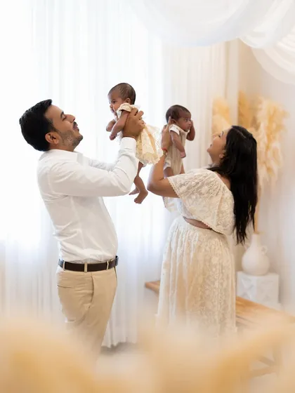 A celebration of new life. In our bright, airy studio, a proud mother and father lift their newborn twins up high, smiling with pure happiness. This pose captures the energy and joy of a new family of four.