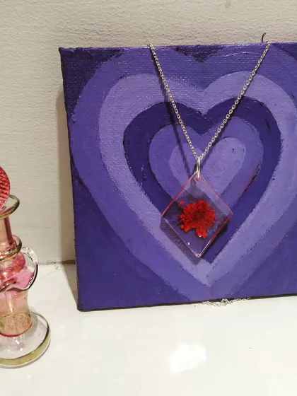 A diamond shaped resin necklace with a small, bright red flower preserved inside. It's displayed against a purple heart painting I made.