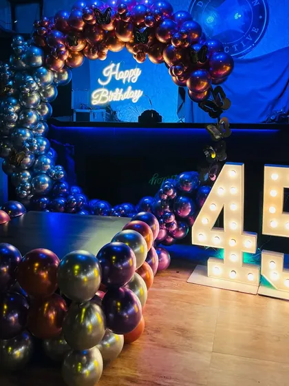 A stylish birthday party backdrop for a 45th birthday. The setup features a circular balloon arch with multi-colored chrome balloons and a 'Happy Birthday' neon sign, complemented by large marquee numbers.