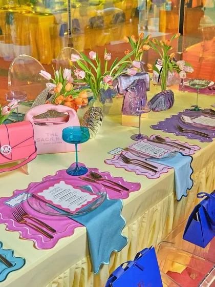 A whimsical and colorful brunch tablescape designed for a couture event. The setting features playful placemats, vibrant glassware, and handbags integrated into the decor, blurring the lines between fashion accessories and table art.
