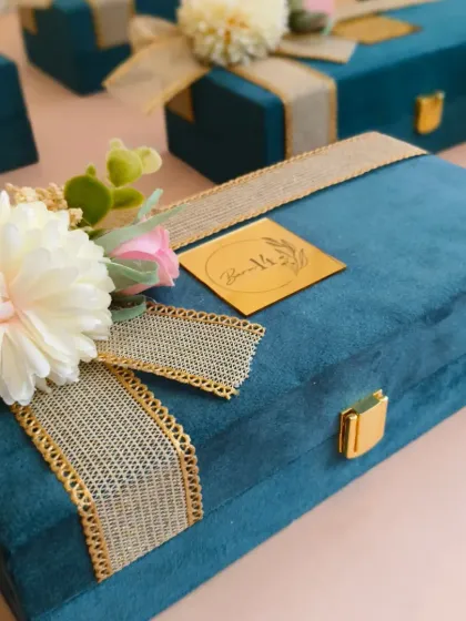A close-up of the exterior of the premium suede box, adorned with a jute-style ribbon and a floral arrangement. The custom gold acrylic logo tag adds a final touch of brand personalization.