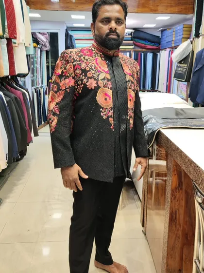 A client trying on a designer black Indo-Western jacket. The piece is adorned with vibrant, multi-colored floral embroidery, making it a showstopper for any festive occasion.